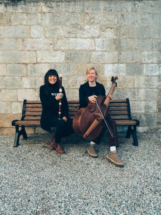 Violeta Motta with her traverso and Anna-Kaisa Meklin with her viola da gamba seated on a bench in front of a stone wall